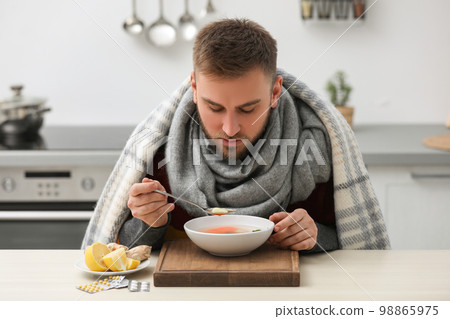 Sick young man eating tasty soup to cure flu at table in kitchen 98865975