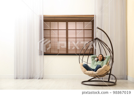 Young woman relaxing in hanging chair near window at home. Space for text 98867019