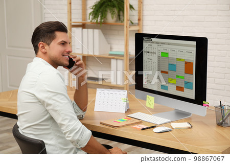 Man talking on smartphone while working with calendar app at table in office 98867067
