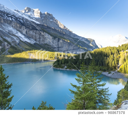 Idyllic morning view of the lake Oeschinensee. Location Swiss alps, Switzerland, Kandersteg district. Blue mountain lake with pine trees and mountains in background. 98867378