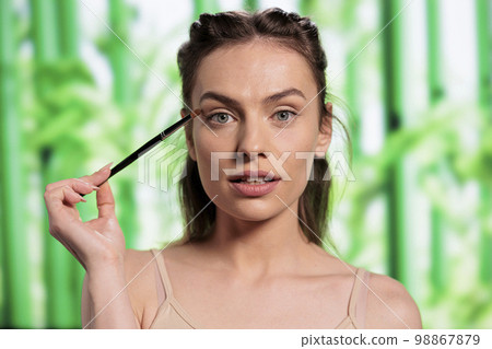 Young beautiful woman doing eyes makeup with brush and looking at camera. Attractive beauty model using natural green decorative cosmetics and applying eyeshadows portrait Young beautiful woman doing eyes makeup with brush and looking at camera. Attractive beauty model using natural green decorative cosmetics and applying eyeshadows portrait 98867879