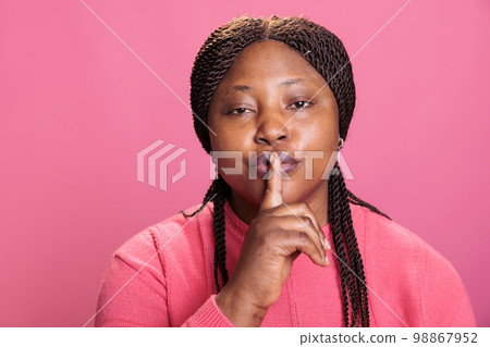 Portrait of serious african american woman making silence gesture by putting forefinger over lips on pink background. Young adult model shushing people indicating secrecy and confidentiality. Portrait of serious african american woman making silence gesture by putting forefinger over lips on pink background. Young adult model shushing people indicating secrecy and confidentiality. 98867952