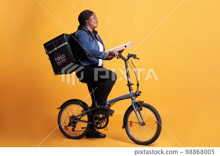 Full body of woman courier holding pizza cardboard while riding bike delivering takeaway meal to client during lunch time. Restaurant worker standing in studio with yellow background Full body of woman courier holding pizza cardboard while riding bike delivering takeaway meal to client during lunch time. Restaurant worker standing in studio with yellow background 98868005
