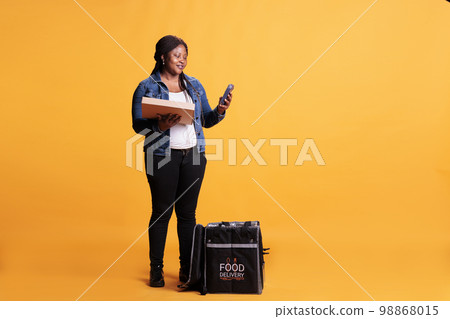 Restaurant employee holding pizza cardboard box while checking customer adreess on phone before delivery order during lunch time. Food transportation service and takeaway concept 98868015