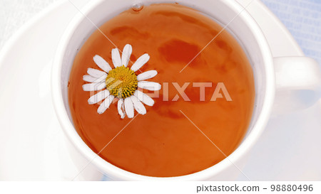 A close-up, a white cup with tea, a daisy flower floating on top of it A close-up, a white cup with tea, a daisy flower floating on top of it 98880496
