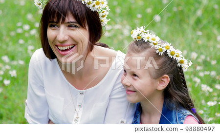 Summer, amidst a chamomile lawn, in the forest, a young woman, a brunette and a girl of seven, mother and daughter weave a wreath of chamomiles, laughing, trying on wreaths 98880497