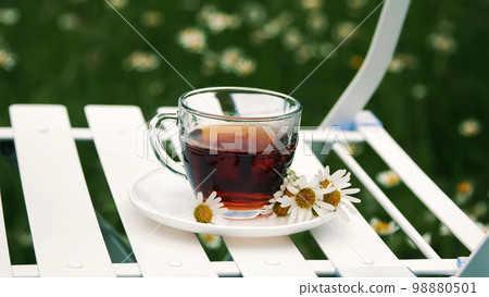 Close-up, deceleration. On a white saucer, decorated with daisies, there is a glass cup with tea. Milk is poured into it. In the background there is a green, chamomile meadow 98880501