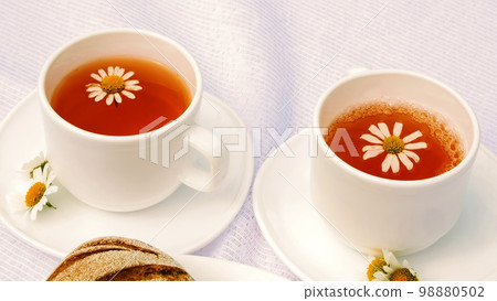 A close-up, a white plate with pastries, biscuits and cookies, decorated with white daisies. On white saucers there are two white cups with chamomile tea A close-up, a white plate with pastries, biscuits and cookies, decorated with white daisies. On white saucers there are two white cups with chamomile tea 98880502