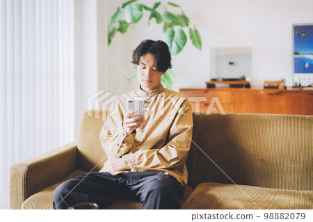 一個男人坐在沙發上，看著他的智能手機 98882079