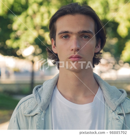 Close up face of an young man is looking to the camera, in the street. Man with calm emotions on his face looks into the camera - outdoors. Handsome guy posing outdoors on green trees background. Close up face of an young man is looking to the camera, in the street. Man with calm emotions on his face looks into the camera - outdoors. Handsome guy posing outdoors on green trees background. 98885702