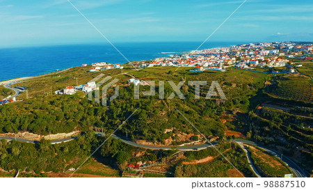Drone view of a beautiful European city with a hilly landscape on ocean background. Aerial view of a small European town against blue sky and Atlantic Ocean.  Beautiful natural landscape. Portugal. 98887510