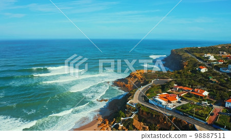 Aerial view of a small European town against blue sky and Atlantic Ocean. Drone view of a beautiful European city with a hilly landscape on ocean background. Beautiful natural landscape. Portugal. 98887511