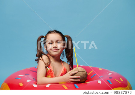 Cute child girl with coconut cocktail smiles looking at camera, posing inside a donut swim ring on blue color background Cute child girl with coconut cocktail smiles looking at camera, posing inside a donut swim ring on blue color background 98887873
