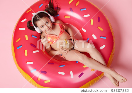 Top view little girl in swimsuit and headphones, lying on inflatable ring, with refreshing coconut drink in hands 98887883