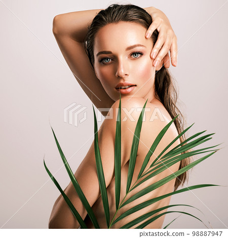 White woman with healthy skin of body and palm leaves. Tanned body of an attractive girl with green plants. Large palm leaves cover the body. 98887947