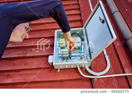 Maintenance of an external electrical panel located on wall of country house, an electrician tightens fixing screw. 98888575