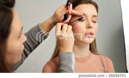 Make-up artist in photo studio doing makeup for beautiful girl. Woman making up using comb for eyelashes. Young woman making makeup in dressing makeup room. Making mua. Applying mascara to the lashes 98888701