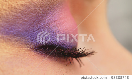 Close-up of a woman's eye with bright shiny makeup.  The blinking eye of a girl in profile. Pink-purple sequined fashion makeup.  Beauty fashion makeup. 98888748