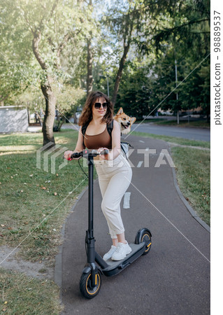 Happy sHappy smiling woman traveler is riding her electro scooter in city parklmiling woman traveler is riding her electro scooter in city parkland with dog Welsh Corgi Pembroke in a special backpack  98889537