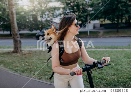 Happy sHappy smiling woman traveler is riding her electro scooter in city parklmiling woman traveler is riding her electro scooter in city parkland with dog Welsh Corgi Pembroke in a special backpack  98889538