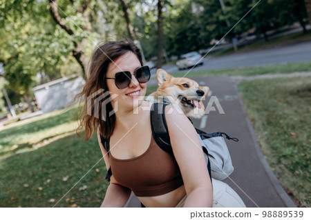 Happy sHappy smiling woman traveler is riding her electro scooter in city parklmiling woman traveler is riding her electro scooter in city parkland with dog Welsh Corgi Pembroke in a special backpack  98889539