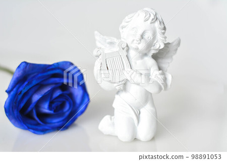 Ceramic figure of an angel on the white glossy surface and a blue rose behind 98891053