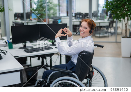 Caucasian woman in wheelchair showing heart sign while sitting in office. Female call center worker. Caucasian woman in wheelchair showing heart sign while sitting in office. Female call center worker. 98891420