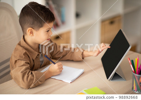 Cheerful small european boy sit at table draws, study uses tablet with blank screen in room interior Cheerful small european boy sit at table draws, study uses tablet with blank screen in room interior 98892668