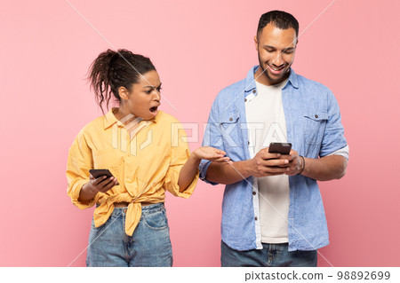 Shocked angry black lady looking at boyfriend's mobile phone, reading his messages, standing together on pink background 98892699