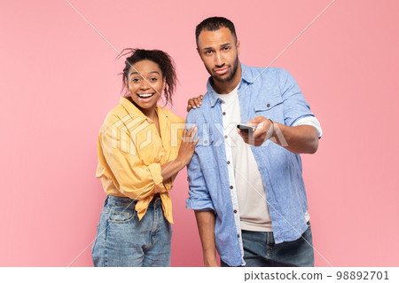 Black couple with remote control watching television program or show on TV over pink studio background 98892701