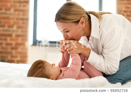 Loving mommy holding her baby daughter hands and kissing, waking up her infant guirl after daytime sleep, lying in bed 98892726