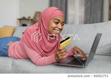 Online shopping. Positive black lady in hijab with laptop and credit card lying on sofa, buying goods in web store 98892751