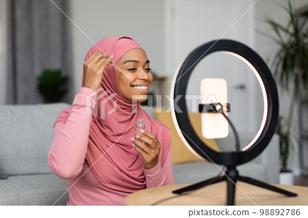 Happy black muslim female beauty blogger making video on smartphone set on ring lamp, showing new facial serum 98892786