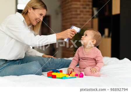 Mother measuring temperature with contactless digital thermometer of her little daughter child, sitting on bed 98892794