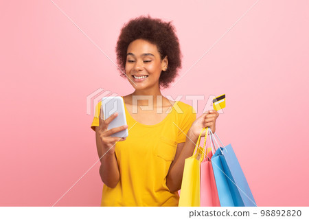 Smiling young black curly lady with many bags of purchases enjoy sale with phone, credit card Smiling young black curly lady with many bags of purchases enjoy sale with phone, credit card 98892820