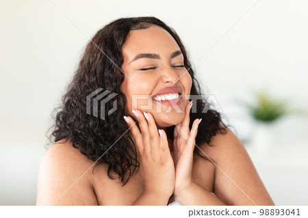 Attractive happy black body positive woman touching her smooth flawless skin on cheeks and smiling with closed eyes Attractive happy black body positive woman touching her smooth flawless skin on cheeks and smiling with closed eyes 98893041