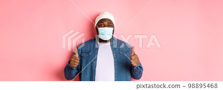 Covid-19, lifestyle and quarantine concept. Satisfied Black man in face mask showing thumbs-up, standing over pink background 98895468