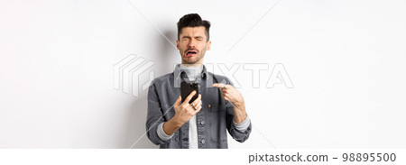 Sad crying guy pointing at smartphone and sobbing, complaining or feeling miserable, standing on white background 98895500