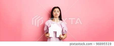 Real estate and insurance concept. Sad frowning asian girl showing paper house cutout and grimacing upset, standing against pink background 98895519