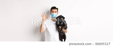Covid-19, animals and quarantine concept. Young man in medical mask holding cute black pug dog, showing okay sign, like and approve, standing over white background 98895527