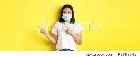 Covid-19, pandemic and social distancing concept. Surprised and happy asian woman in medical mask, showing advertisement, pointing right and smiling, yellow background Covid-19, pandemic and social distancing concept. Surprised and happy asian woman in medical mask, showing advertisement, pointing right and smiling, yellow background 98895548