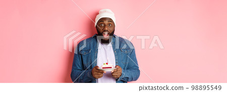 Excited Black man celebrating birthday, staring at camera amazed while making wish on bday cake with candle, standing over pink background Excited Black man celebrating birthday, staring at camera amazed while making wish on bday cake with candle, standing over pink background 98895549