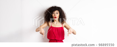Impressed woman in red dress say wow, showing promo deal, pointing fingers down with amazed face, standing over white background Impressed woman in red dress say wow, showing promo deal, pointing fingers down with amazed face, standing over white background 98895550