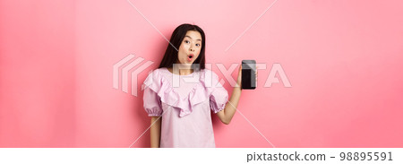 Online shopping. Excited asian woman say wow, showing empty smartphone screen and looking amazed at camera, standing against pink background Online shopping. Excited asian woman say wow, showing empty smartphone screen and looking amazed at camera, standing against pink background 98895591