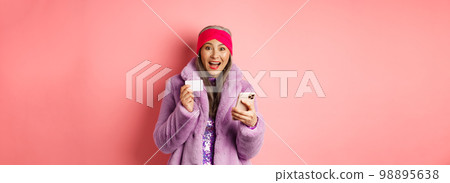 Online shopping and fashion concept. Happy asian female checking out internet promotions, holding credit card and smartphone, pink background 98895638