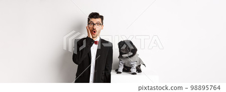 Animals, party and celebration concept. Shocked handsome man in formal suit and cute dog in costume, staring at camera amazed, standing over white background Animals, party and celebration concept. Shocked handsome man in formal suit and cute dog in costume, staring at camera amazed, standing over white background 98895764
