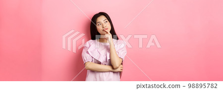 Pensive smiling asian girl making choice, looking at upper left corner logo and thinking, having an idea, standing cunning on pink background Pensive smiling asian girl making choice, looking at upper left corner logo and thinking, having an idea, standing cunning on pink background 98895782