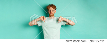 Disappointed caucasian guy with red messy hair and glasses showing thumbs down, dislike something, frowning upset and displeased, standing over mint background Disappointed caucasian guy with red messy hair and glasses showing thumbs down, dislike something, frowning upset and displeased, standing over mint background 98895794