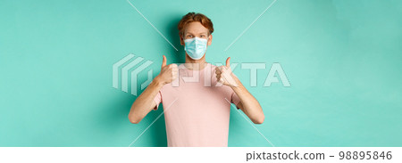 Covid-19, pandemic and lifestyle concept. Cheerful redhead guy in medical mask showing thumbs up in approval, like and praise product, standing over turquoise background 98895846