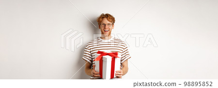 Valentines day and holidays concept. Cheerful young man holding gift box and smiling grateful, receiving presents, standing over white background Valentines day and holidays concept. Cheerful young man holding gift box and smiling grateful, receiving presents, standing over white background 98895852